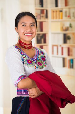 Geleneksel Andean bluzu giyen güzel genç bir avukat elinde kırmızı bir ceket kameraya gülümsüyor, arka planda kitaplıklar var.