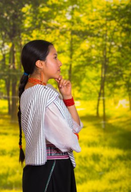 Geleneksel giyen güzel genç kadın and bluz ile kolye, kamera, düşünceli beden dili ve yüz ifadesi için poz ayakta, yeşil orman arka plan