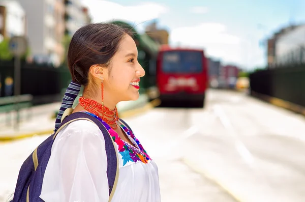 Bella giovane donna che indossa camicetta andina tradizionale e zaino blu, in attesa di autobus sulla piattaforma della stazione all'aperto, sorridendo felicemente, come si vede dall'angolazione del profilo — Foto Stock