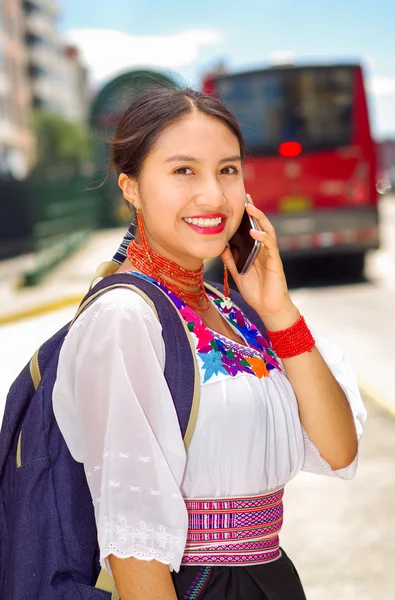 Mujer joven y bonita con blusa andina tradicional y mochila azul, esperando el autobús en la plataforma de la estación al aire libre, hablando por teléfono sonriendo felizmente, como se ve desde el ángulo del perfil — Foto de Stock