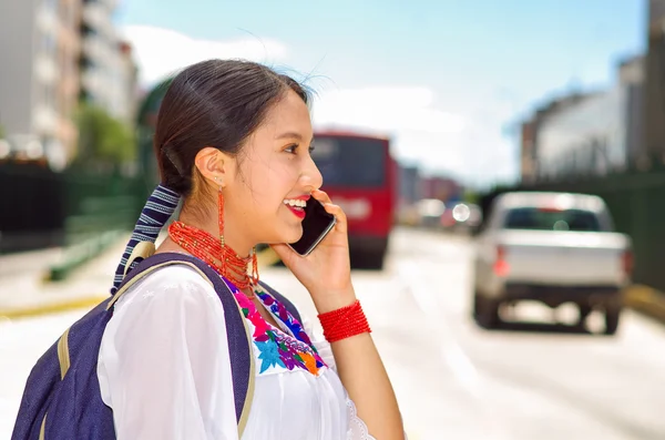 Hübsche junge Frau in traditioneller Anden-Bluse und blauem Rucksack, die am Bahnsteig auf den Bus wartet, am Telefon glücklich lächelnd, aus dem Blickwinkel des Profils gesehen — Stockfoto