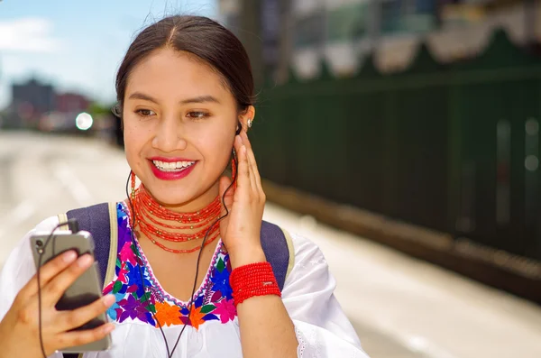 Mujer bastante joven con blusa andina tradicional y mochila azul, esperando el autobús en la plataforma de la estación al aire libre, mirando a la pantalla móvil con auriculares conectados sonriendo felizmente —  Fotos de Stock