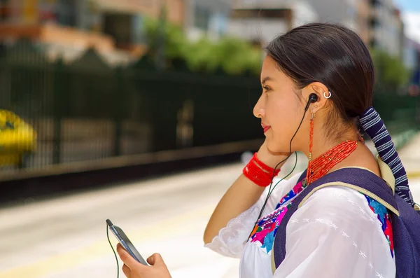 漂亮的年轻女子穿着传统的上衣和蓝色背包，在户外站台等车，用手机听音乐，用耳机连接，开心地微笑 — 图库照片