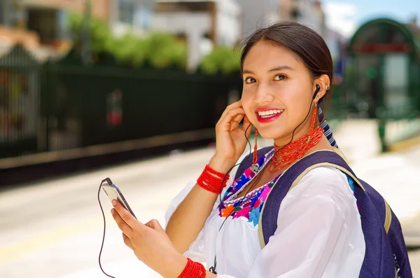 Pretty young woman wearing traditional andean blouse and blue backpack, waiting for bus at outdoors station platform, using mobile phone listening to music with headphones connected, smiling happily — Stock Photo, Image