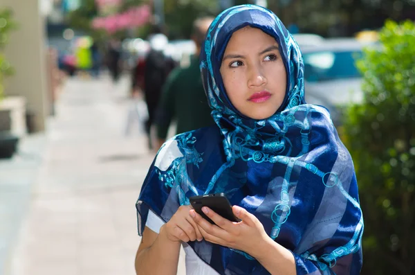 Bella giovane donna musulmana indossa hijab di colore blu, utilizzando il telefono cellulare in background urbano all'aperto — Foto Stock