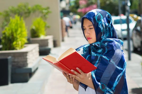 Piękna młoda muzułmańska kobieta ubrana w niebieskie, kolorowe hidżab, posiadająca grubą księgę trzciny i czytając na ulicy, na zewnątrz tło miejskie — Zdjęcie stockowe