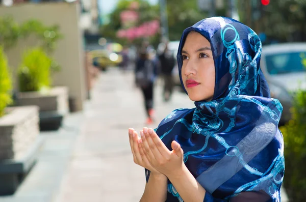 Bella giovane donna musulmana che indossa hijab di colore blu, mani pieghevoli che eseguono la preghiera, all'aperto sfondo urbano — Foto Stock