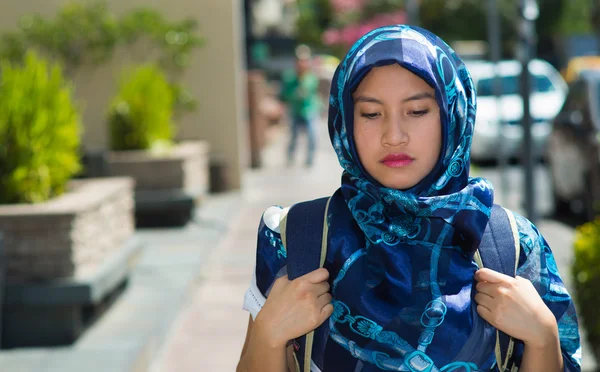 Mooie jonge moslim vrouw dragen blauw gekleurde hijab en rugzak, poseren met doordachte ernstige gezichtsuitdrukking in de straat, buitenshuis stedelijke achtergrond — Stockfoto