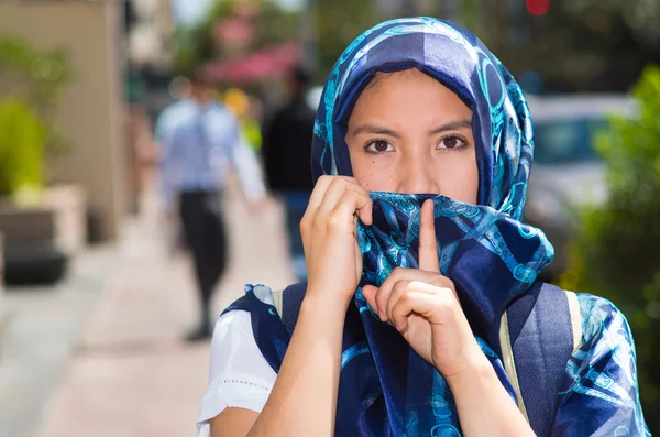 Mooie jonge moslim vrouw dragen blauw gekleurde hijab en rugzak, poseren terwijl het bedekken van de helft van gezicht het maken van stilte symbool met behulp van vinger over lippen, buitenshuis stedelijke achtergrond — Stockfoto