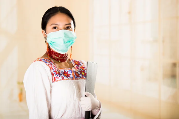 Mujer hermosa joven vestida con abrigo de médicos y collar rojo, cara cubierta con máscara facial mirando a la cámara, fondo de clínica clara de huevo — Foto de Stock