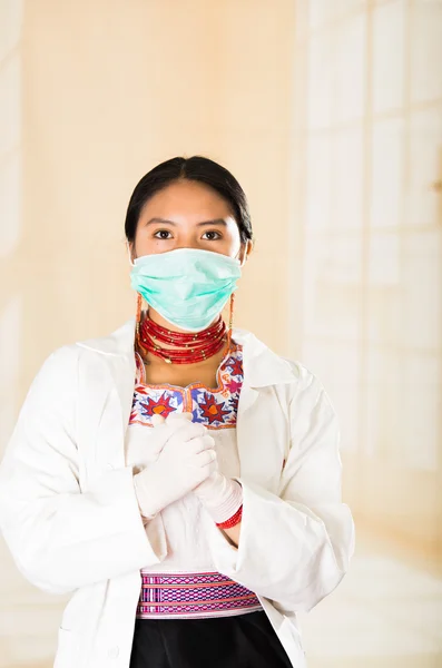 Mujer hermosa joven vestida con abrigo de médicos y collar rojo, cara cubierta con máscara facial mirando a la cámara, fondo de clínica clara de huevo — Foto de Stock