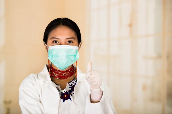 Jeune belle femme vêtue d'un manteau de médecin et d'un collier rouge, visage couvert d'un masque facial regardant vers la caméra, fond de clinique blanc d'oeuf — Photo