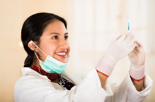 Young beautiful woman dressed in doctors coat and red necklace, facial mask pulled down to chin, holding up syringe smiling happily, egg white clinic background — Stock Photo, Image