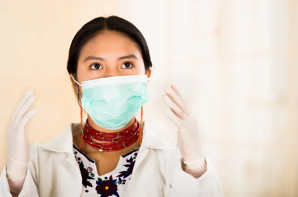 Mujer hermosa joven vestida con abrigo de médicos y collar rojo, cara cubierta con máscara facial mirando a la cámara, fondo de clínica clara de huevo — Foto de Stock