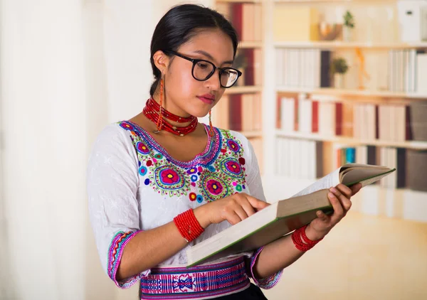 Beautiful young lawyer wearing traditional andean blouse and glasses, holding book reading, bookshelves background