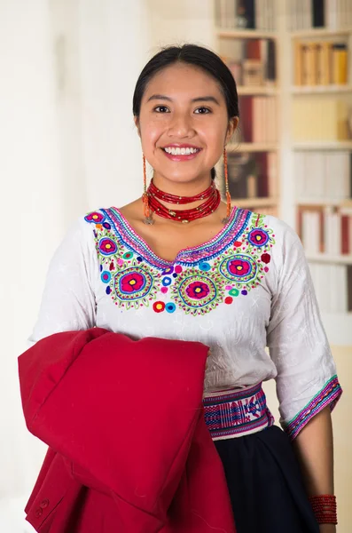 Vacker ung advokat bär traditionella andean blus, håller röd jacka ler mot kameran, bokhyllor bakgrund — Stockfoto