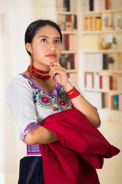 Vacker ung advokat bär traditionella andean blus, håller röd jacka ler mot kameran, bokhyllor bakgrund — Stockfoto