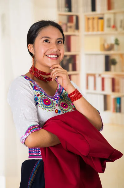 Geleneksel Andean bluzu giyen güzel genç bir avukat elinde kırmızı bir ceket kameraya gülümsüyor, arka planda kitaplıklar var. — Stok fotoğraf