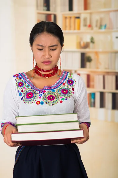 Beautiful young lawyer wearing traditional andean blouse and red necklace, holding stack of books, upset facial expression, bookshelves background — Stockfoto