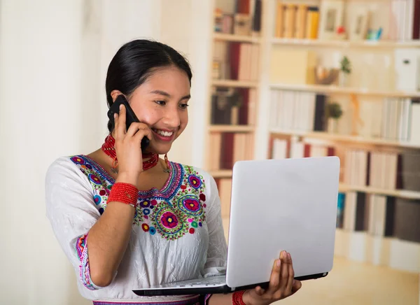 Belle jeune avocate portant un chemisier andin traditionnel et un collier rouge, tenant un ordinateur portable parlant sur un téléphone souriant, fond de bibliothèque — Photo