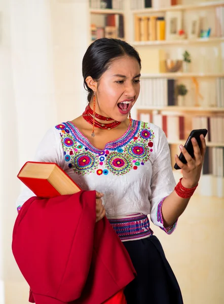 Bela jovem advogado vestindo blusa andina tradicional com colar, segurando jaqueta vermelha e livro ao usar o telefone móvel, olhando chocado na tela, prateleiras de fundo — Fotografia de Stock