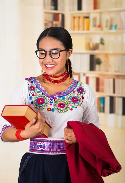 Piękny młody prawnik ubrany w tradycyjną andyjską bluzkę, okulary i naszyjnik, trzymający czerwoną kurtkę jedną ręką, grubą książkę w drugiej, uśmiechnięty szczęśliwie, tło półek na książki — Zdjęcie stockowe