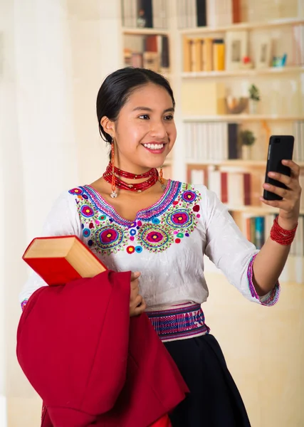 Beautiful young lawyer wearing traditional andean blouse with necklace, holding red jacket and book while using mobile phone, smiling happily, bookshelves background — Stock fotografie