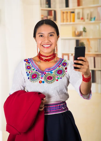 Vacker ung advokat bär traditionella andean blus och halsband, hålla röd jacka när du använder mobiltelefon, ler glatt, bokhyllor bakgrund — Stockfoto