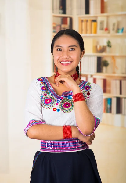Beautiful young lawyer wearing black skirt, traditional andean blouse with necklace, standing posing for camera, smiling happily, bookshelves background
