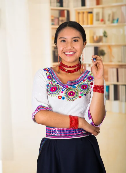 Vacker ung advokat bär svart kjol, traditionell andean blus med halsband, stående poserar för kameran, ler glatt, bokhyllor bakgrund — Stockfoto