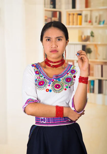 Hermosa abogada joven con falda negra, blusa andina tradicional con collar, posando para cámara, expresión facial seria, fondo de estanterías —  Fotos de Stock