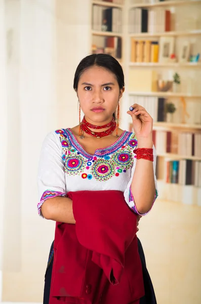 Beautiful young lawyer wearing black skirt, traditional andean blouse with necklace, standing posing for camera, holding red jacket, serious facial expression, bookshelves background