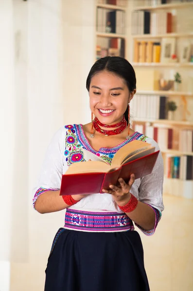 Bella giovane avvocato indossa gonna nera, camicetta andina tradizionale con collana, in piedi in posa per la fotocamera, tenendo lettura del libro rosso felicemente, scaffali sfondo . — Foto Stock