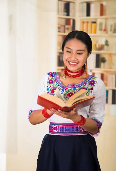 Bella giovane avvocato indossa gonna nera, camicetta andina tradizionale con collana, in piedi in posa per la fotocamera, tenendo lettura del libro rosso felicemente, scaffali sfondo . — Foto Stock