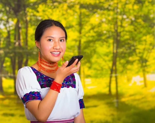 Bella giovane donna indossa camicetta andina tradizionale con collana, in piedi in posa per la fotocamera, tenendo il telefono cellulare parlando, sfondo verde foresta — Foto Stock
