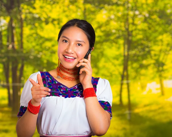Beautiful young woman wearing traditional andean blouse with necklace, standing posing for camera, holding mobile phone talking, green forest background — Stock Photo, Image