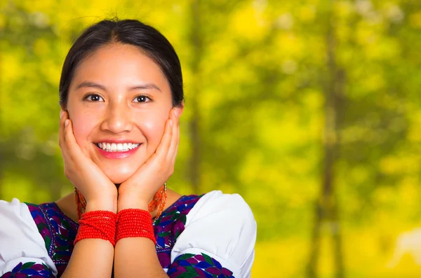 Headshot piękna młoda kobieta ubrana w tradycyjny Andyjskiej bluzki z czerwonym naszyjnikiem, stwarzając dla kamery dotykając twarzy za pomocą rąk, podczas gdy uśmiechnięta szczęśliwie, zielone tło lasu — Zdjęcie stockowe