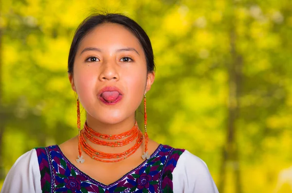Cabeza hermosa mujer joven con blusa andina tradicional con collar rojo, posando para la cámara con la lengua hacia fuera, fondo verde bosque —  Fotos de Stock