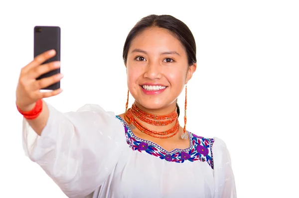 Hermosa joven de pie vistiendo una blusa andina tradicional y un collar rojo, sosteniendo el teléfono móvil tomando selfie mientras sonríe felizmente, fondo blanco del estudio — Foto de Stock