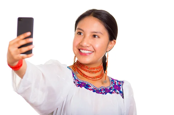 Hermosa joven de pie vistiendo una blusa andina tradicional y un collar rojo, sosteniendo el teléfono móvil tomando selfie mientras sonríe felizmente, fondo blanco del estudio — Foto de Stock