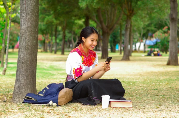 Mujer joven vestida con falda y blusa andina tradicional con collar rojo a juego, sentada en la hierba junto al árbol en el área del parque, enviando mensajes de texto usando el teléfono móvil mientras sonríe felizmente —  Fotos de Stock