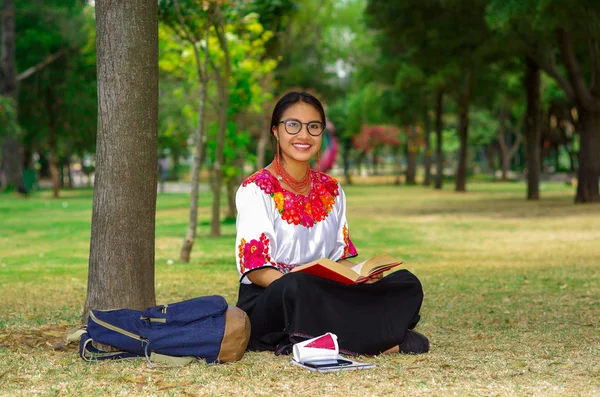 Jonge vrouw met traditionele bril, Andes rok en blouse met bijpassende rode ketting, zittend op gras naast de boom in het park, ontspannen tijdens het lezen boek, vrolijk glimlachen — Stockfoto