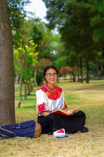 Mulher jovem vestindo óculos tradicionais, saia andina e blusa com colar vermelho combinando, sentado na grama ao lado da árvore na área do parque, relaxante enquanto lê o livro, sorrindo feliz — Fotografia de Stock