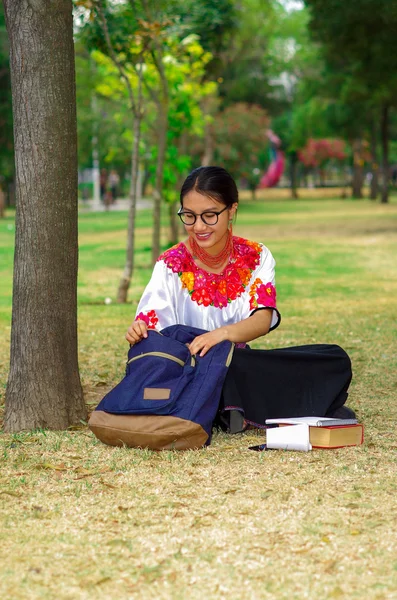 Jovem mulher vestindo saia e blusa andina tradicional com colar vermelho combinando, sentado na grama ao lado da árvore na área do parque, relaxando enquanto olha para a mochila, sorrindo feliz — Fotografia de Stock