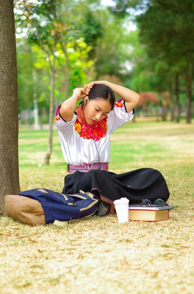 Junge Frau in traditionellem Anden-Rock und Bluse mit passender roter Halskette, sitzt auf dem Rasen neben einem Baum im Parkbereich und entspannt beim Haaranlegen — Stockfoto