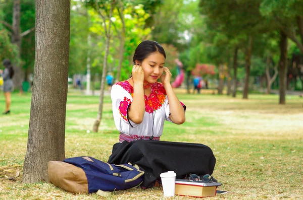 Geleneksel andean etek ve bluz giyen genç bir kadın, kırmızı kolyeli, park alanındaki ağacın yanındaki çimlere oturmuş, cep telefonuyla bağlanmış, mutlu bir şekilde gülümsüyor. — Stok fotoğraf