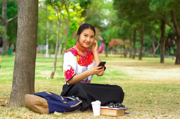 Ung kvinna bär traditionell andean kjol och blus med matchande rött halsband, sitter på gräs bredvid träd i parkområde, koppla av medan du använder mobiltelefon hörlurar anslutna, ler glatt — Stockfoto