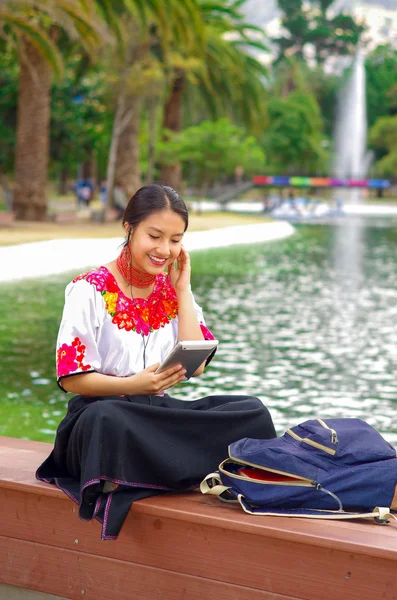 Giovane donna che indossa gonna tradizionale andina e camicetta con collana rossa abbinata, seduta sulla panchina accanto al lago nella zona del parco, rilassante mentre si utilizza tablet, sorridente felicemente — Foto Stock