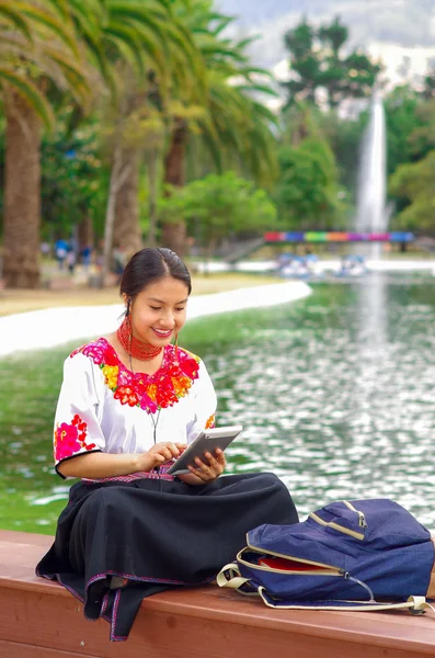 Jonge vrouw in traditionele Andes rok en blouse met bijpassende rode ketting, zittend op een bankje naast het meer in het park, ontspannen tijdens het gebruik van tablet, vrolijk glimlachen — Stockfoto