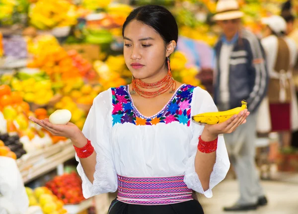 Beautiful young hispanic woman wearing andean traditional blouse posing for camera holding banana and onion inside fruit market, colorful healthy food selection in background